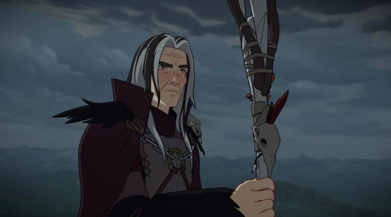 An animated elderly character with long gray hair, dressed in dark medieval attire, holding a staff adorned with feathers and a skull, set against a dramatic, cloudy sky and mountainous backdrop.